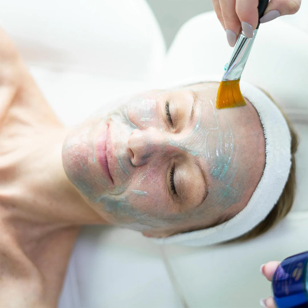 Person lying down with eyes closed while a brush applies a gray-blue facial mask to their face.