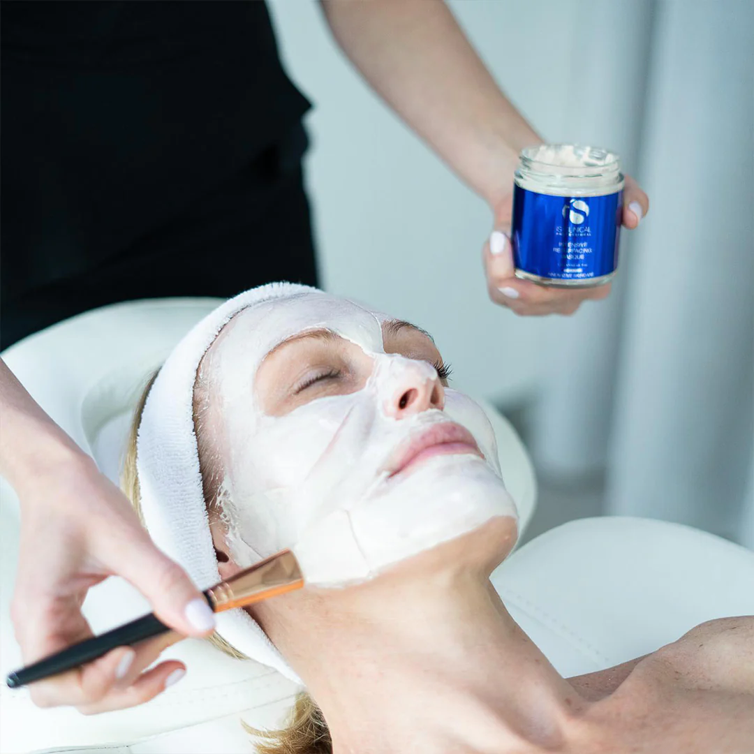 Person receiving facial treatment with white mask applied and blue jar product held nearby.