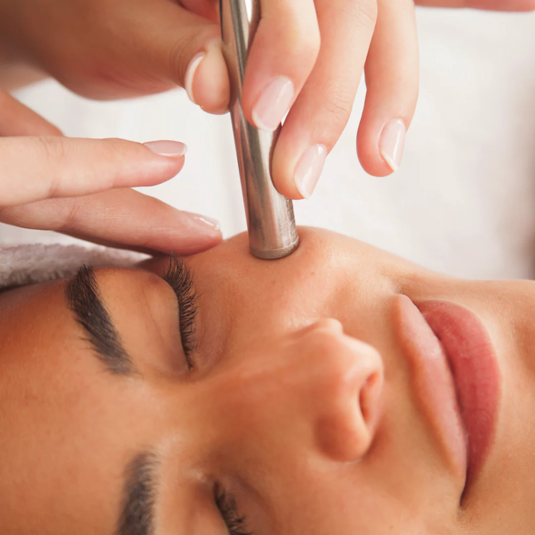 Metal facial treatment device held above person's cheek during skincare procedure.