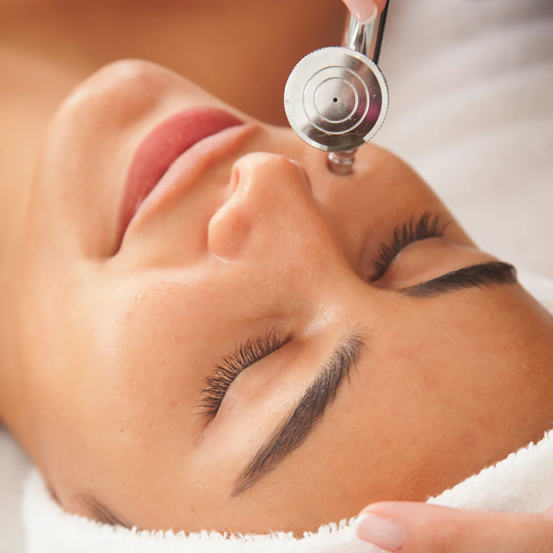 Handheld facial treatment device with circular metallic head being used on person's face during skincare procedure.